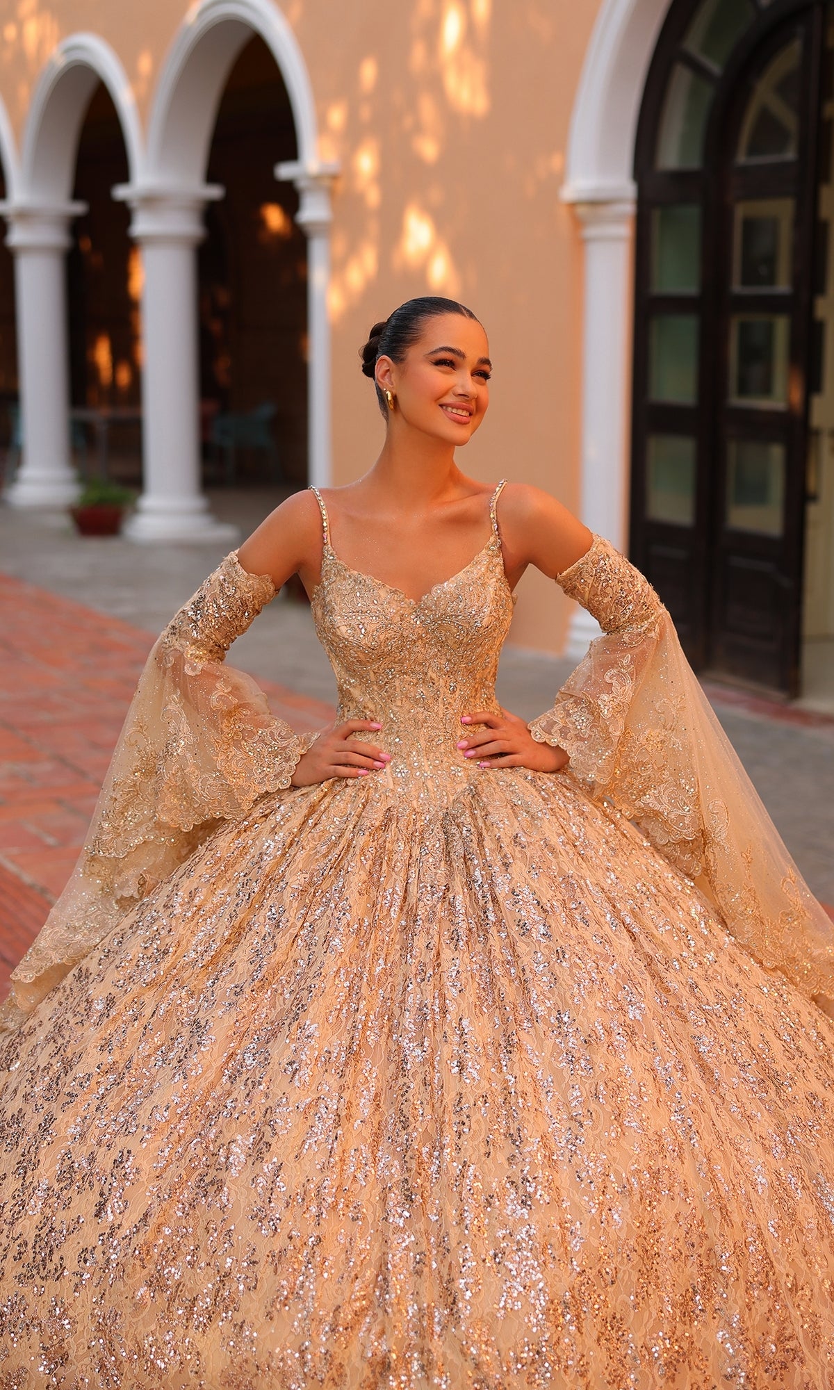 A woman in the Quinceanera Dress 54303 by Amarra, a sparkling gold sequin ball gown, poses outdoors in front of an arched building, smiling with her hands on her hips.